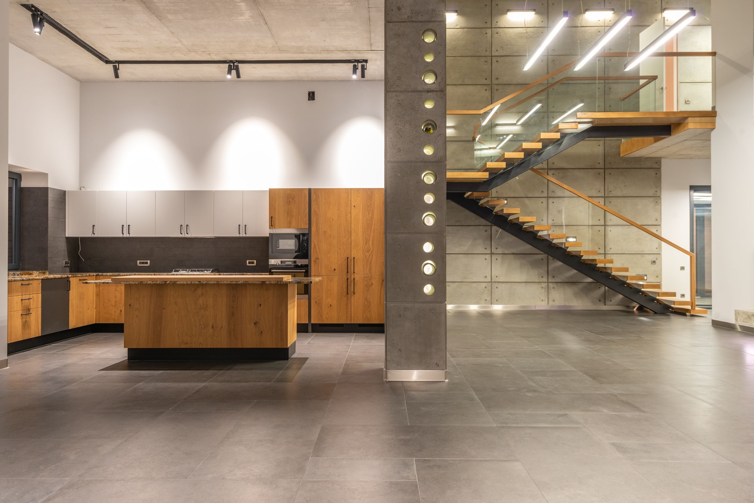 Zona de día de una casa reformada con cocina con isla de madera y escalera de peldaños de madera junto a un muro de hormigón