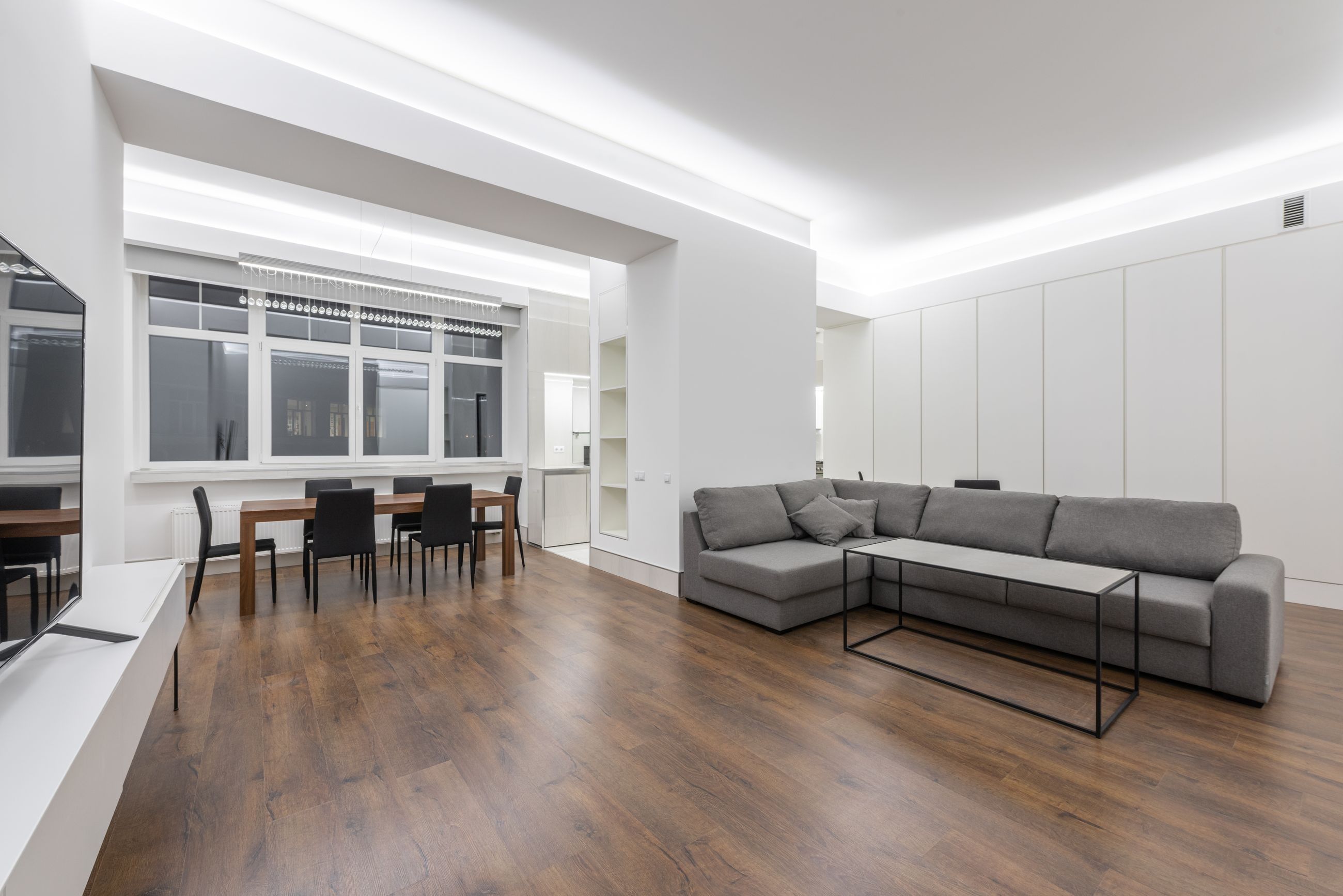 Salón renovado con techo de luz indirecta, paredes blancas, suelo de madera y sofá gris